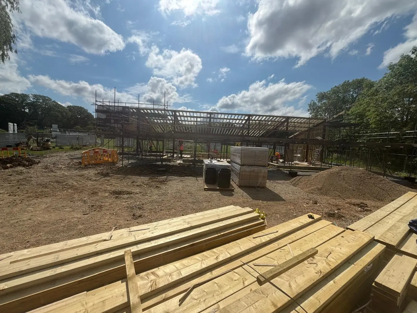 A wide view of the pavilion site, with the steel frame and timber roof taking shape behind stacked joists